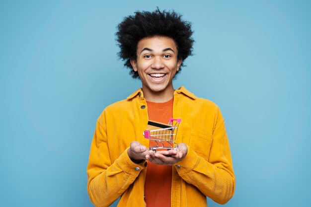 Smiling African American man holding shopping trolley with credit card, isolated. Shopping, sales