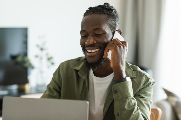 Smiling african american man talking on smartphone and looking at laptop working at home free space