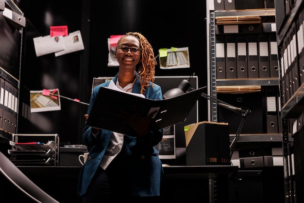 Smiling african american woman detective holding case folder portrait. Young police inspector secretary reading investigation file and looking at camera in office room at night time