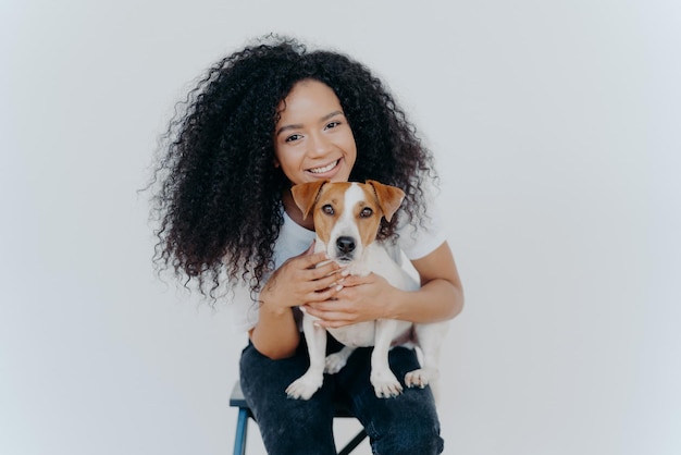 Smiling African American woman poses with domestic animal, looks gladfully at camera, cuddles dog, sits on chair, isolated over white background. Two family members. Homely relax. Forever together