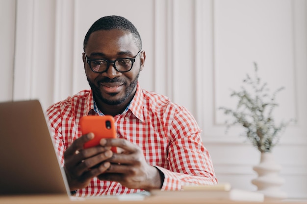 Smiling African ethnicity man office worker sitting at desk holding smartphone chatting with friends