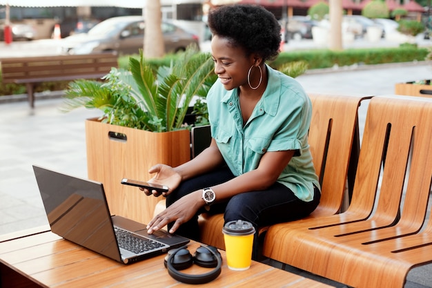 Smiling African girl sitting in a street cafe and working at the computer. The girl checks the data on the laptop and in the phone. Work online, business online.