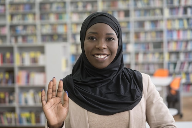 Smiling African woman wearing hijab using laptop computer having video call sitting in library