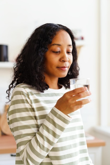 Smiling africanamerican female looking at glass of clear water on soft focused background