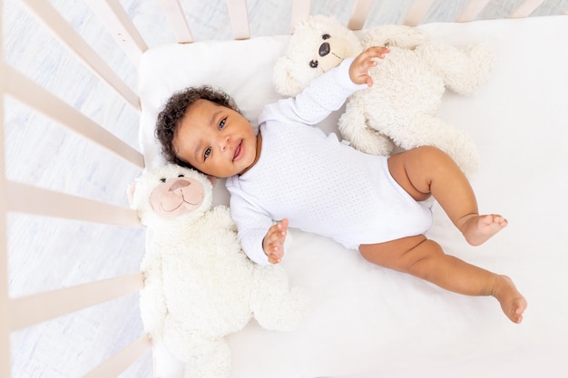 Smiling AfricanAmerican little baby in white sleeping bed with bear toys