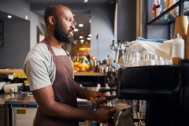 Smiling Afro American man in apron holding white cup under tap of coffee machine while making espresso