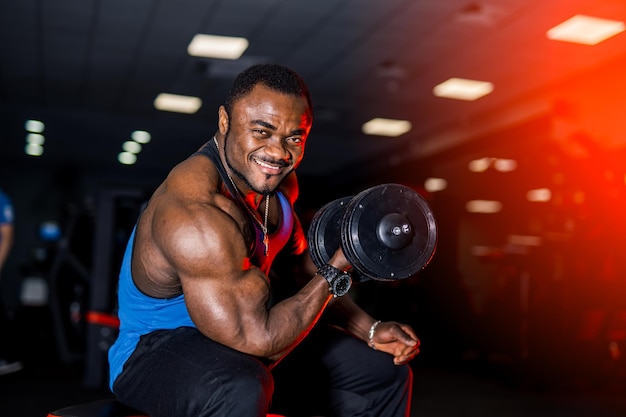 Smiling afro american sports man looking at camera over black background with dumbbell in hand Perfect muscular body with strong muscles