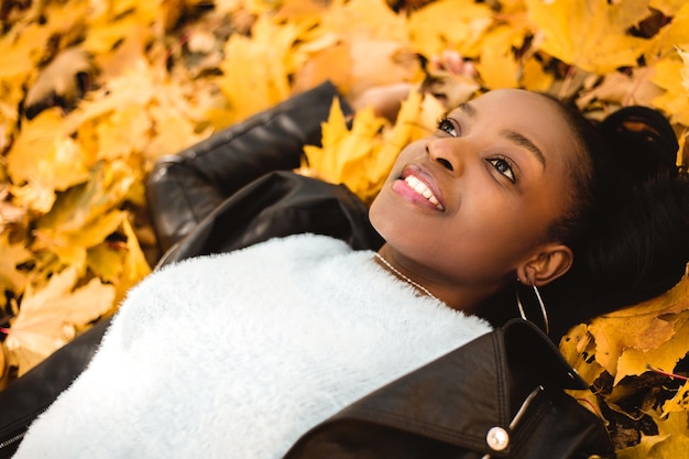 Smiling amazed african american woman lying on the ground with pile of yellow leaves