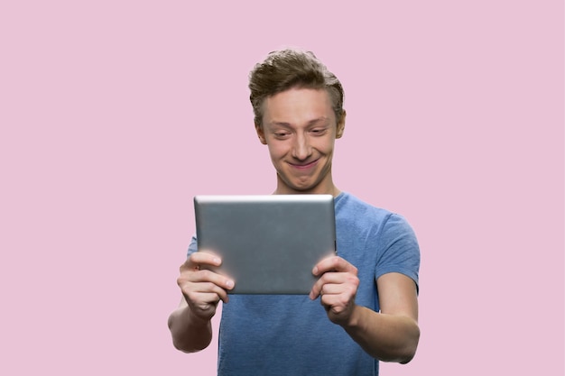 Smiling american guy looking at the tablet. Caucasian schoolboy isolated on pink background.