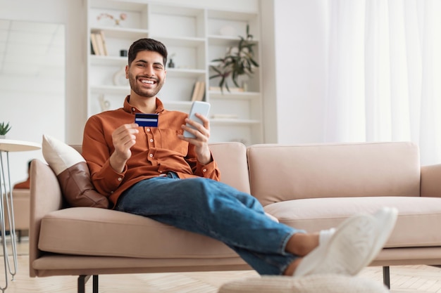 Smiling arab guy using phone and credit card at home