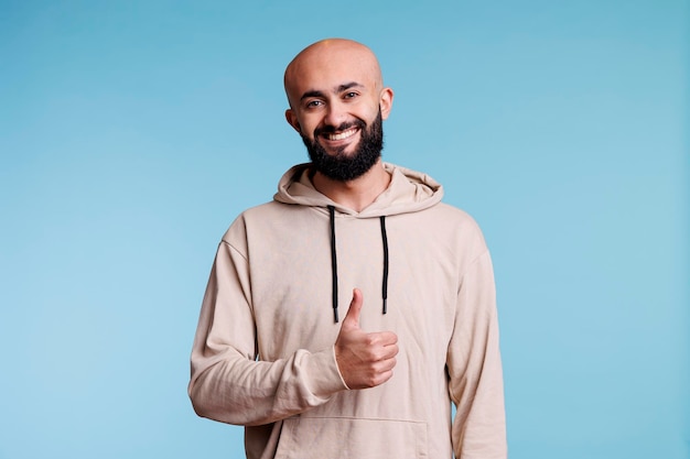 Smiling arab man with thumb up showing approval and looking at camera. Cheerful young person with carefree facial expression posing with satisfaction and agreement gesture studio portrait