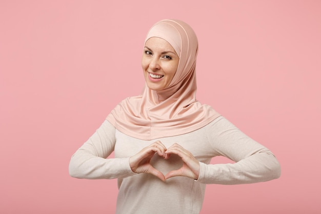 Smiling arabian muslim woman in hijab light clothes posing isolated on pink background. People religious Islam lifestyle concept. Mock up copy space. Showing shape heart with hands, heart-shape sign.