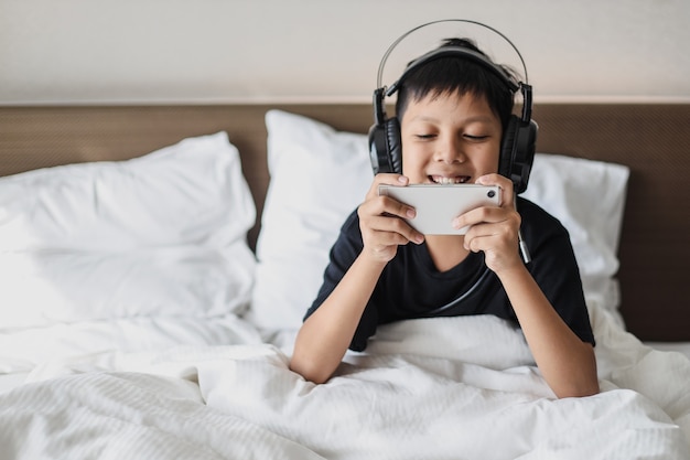 Smiling asian boy in headphone sitting on the bed while playing game on a smart phone