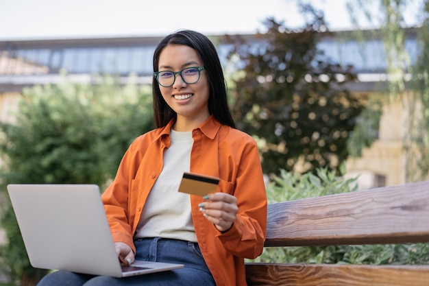 Smiling asian business woman holding credit card using laptop computer shopping online