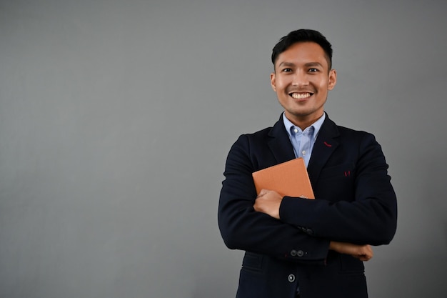 Smiling Asian businessman in formal business suit with a book isolated grey background