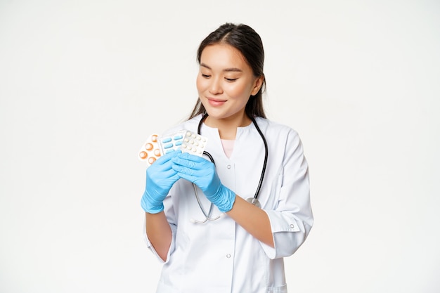 Smiling asian doctor showing medical pills, advertising vitamins, holding drugs with sterile gloves, wearing medical uniform, white background.