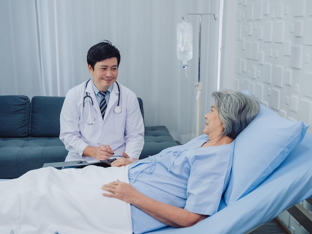 Smiling Asian doctor in white suit take notes while discussing happy Asian elderly senior woman patient who lying on bed with receiving saline solution in hospital Health care and medical concept