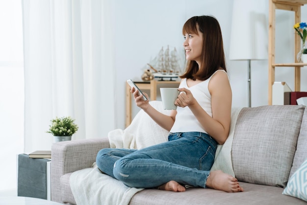 smiling asian female is sitting leisurely in living room enjoying her morining coffee. chinese pretty woman holidng phone and mug is gazing out the window pondering.