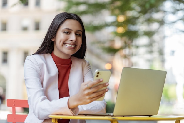 smiling asian freelancer using laptop holding mobile phone working online sitting at workplace