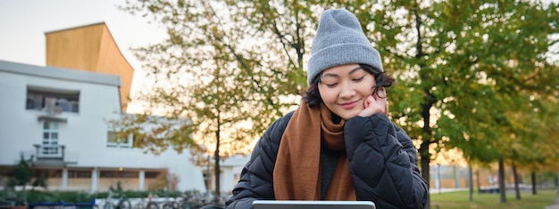 Smiling asian girl student sits on bench in park alone reading using social media app on digital