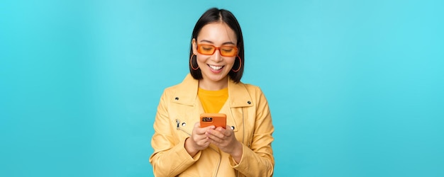 Smiling asian girl in sunglasses using smartphone app holding mobile phone standing over blue background Copy space