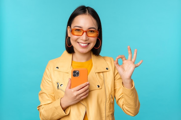 Smiling asian girl with smartphone shwoing okay ok sign in approval standing over blue background