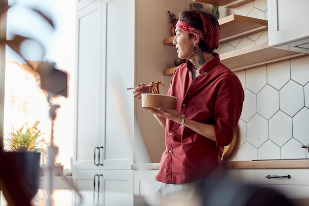 Smiling asian guy leaning on the kitchen countertop and eating at home