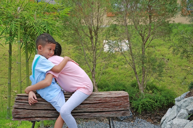 Smiling Asian little brother and young sister embracing while sit on wooden chair in the park