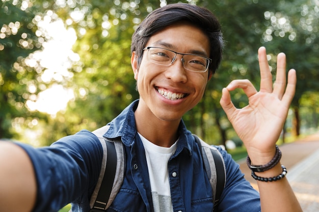 Smiling asian male student in eyeglasses making selfie and showing ok sign while looking at the camera outdoors