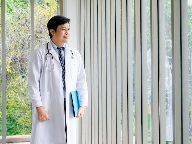 Smiling Asian man doctor portrait standing near the glass windows in medical office with green view Confident Asian adult male doctor with stethoscope holding blue document folder looking outside