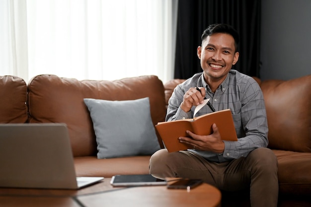 Smiling Asian man making list or taking notes on his notebook while sitting on sofa