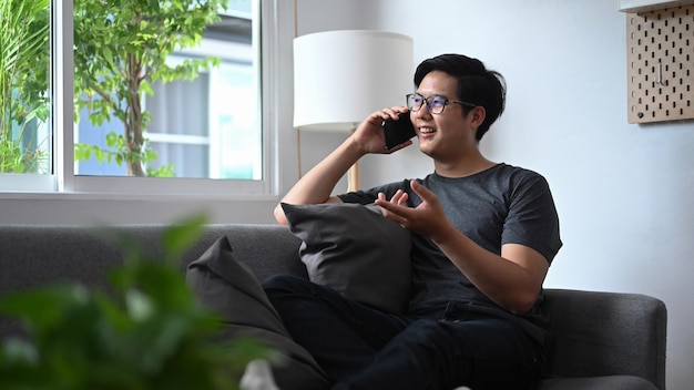 Smiling Asian man talking on smart phone and sitting on sofa in living room