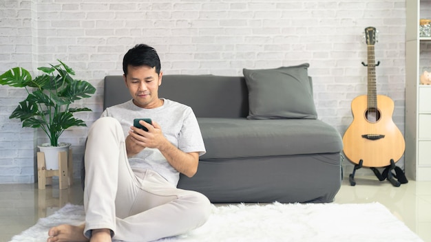 Smiling asian man wearing casual clothes sitting on a couch at the living room. Asian man using mobile phone chatting in living room.