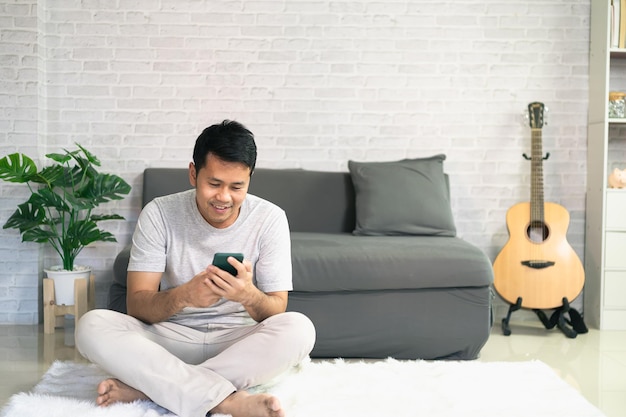 Smiling asian man wearing casual clothes sitting on a couch at the living room. Asian man using mobile phone chatting in living room.