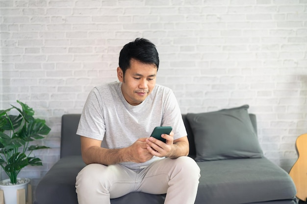 Smiling asian man wearing casual clothes sitting on a couch at the living room Asian man using mobile phone chatting in living room