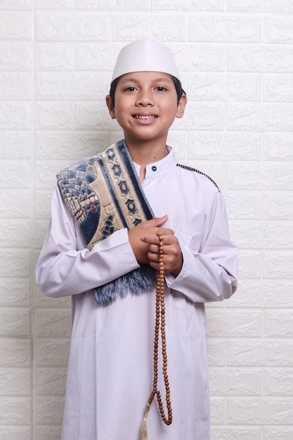 Smiling Asian muslim kid wearing white rob and skullcap holding prayer beads, prayer mat on shoulder