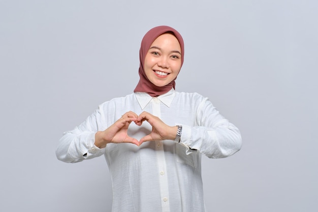 Smiling Asian Muslim woman showing heart gesture making love sign with fingers isolated over white background