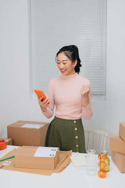 Smiling Asian online business owner prepares parcels and check online orders for delivery to customers on her smartphone