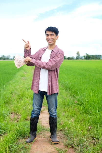 Smiling Asian peasant man holding banknotes and pointing his finger in the middle of the rice field