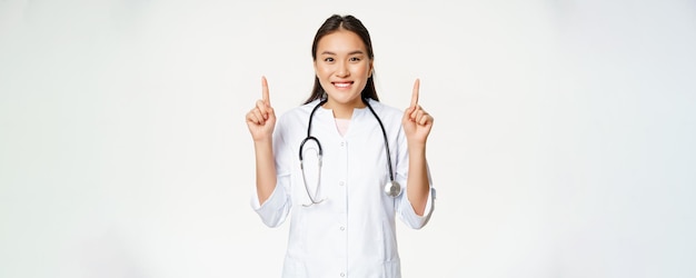 Smiling asian woman doctor wearing medical robe pointing fingers up at advertisement showing promo p