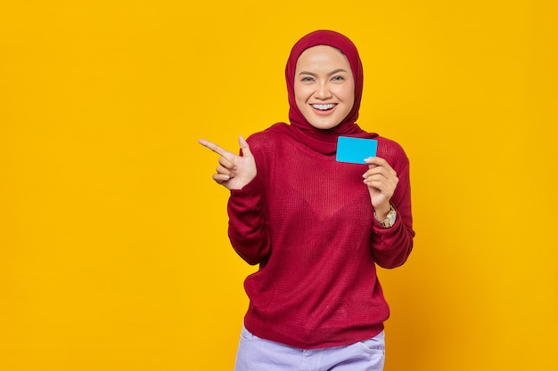 Smiling Asian woman holding a credit card while pointing finger at copy space on yellow background