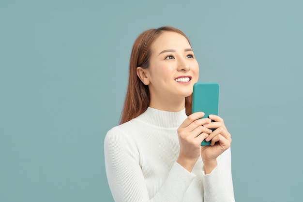 Smiling asian woman holding smartphone and looking away over violet wall