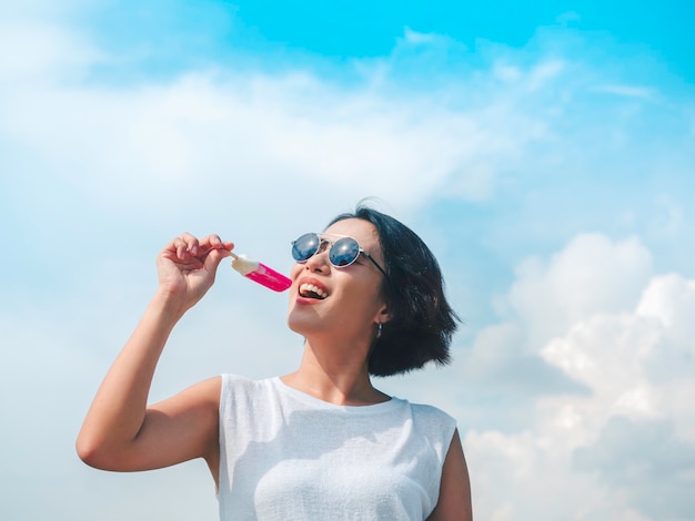 Smiling Asian woman short hair in casual white sleeveless shirt wearing sunglasses holding pink popsicle on blue sky background in summertime. Women eating popsicles, freshy summer season concept.