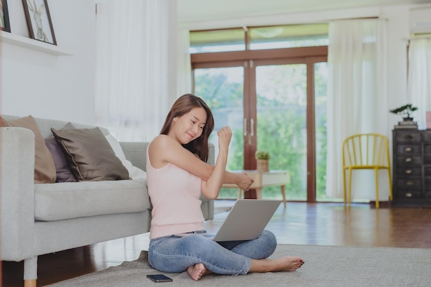 Smiling Asian woman stretching her shoulder for relax after working from home via laptop