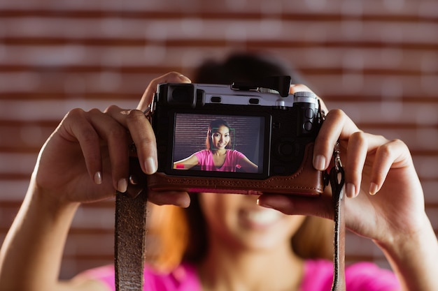 Smiling asian woman taking picture with camera on brick wall