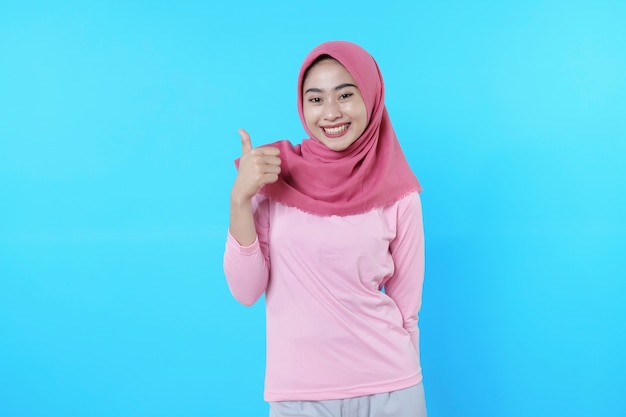 Smiling asian woman with give a thumb isolated on light blue background wearing hijab with pink t shirt