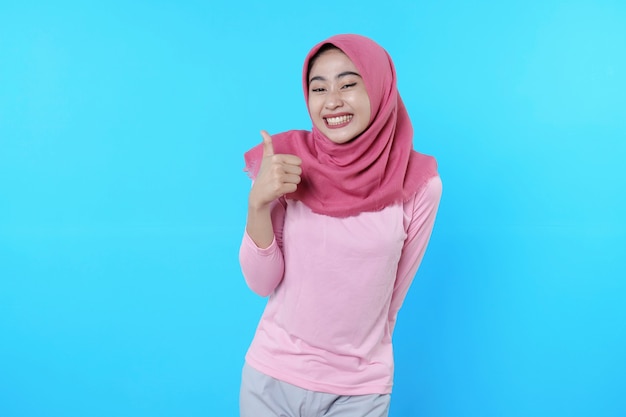 Smiling asian woman with give a thumb isolated on light blue background wearing hijab with pink t shirt