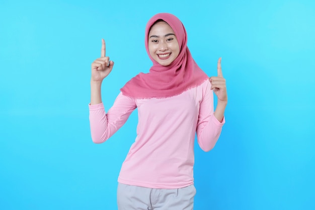 Smiling asian woman with her finger pointing isolated on light blue background wearing hijab with pink t shirt