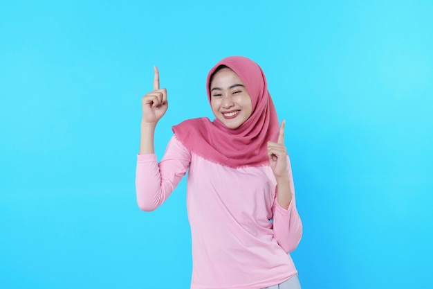 Smiling asian woman with her finger pointing isolated on light blue background wearing hijab with pink t shirt