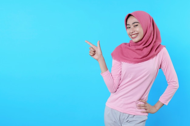 Smiling asian woman with her finger pointing isolated on light blue background wearing hijab with pink t shirt